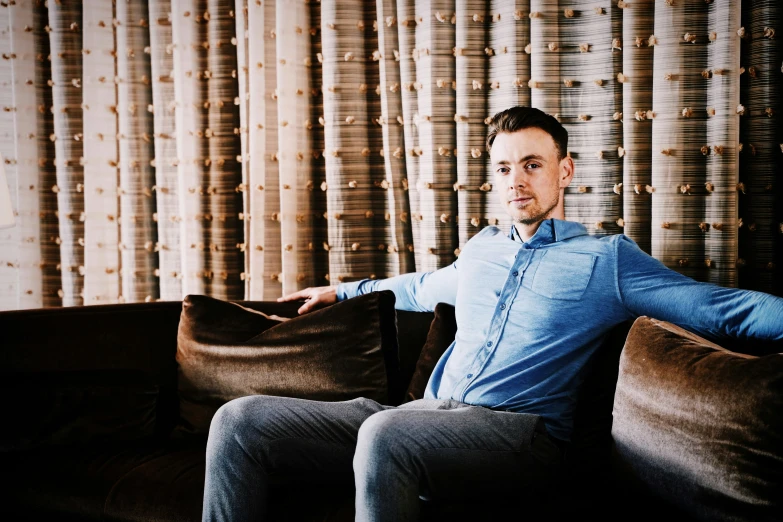 man sitting on couch with arm extended out, by a window of tan color