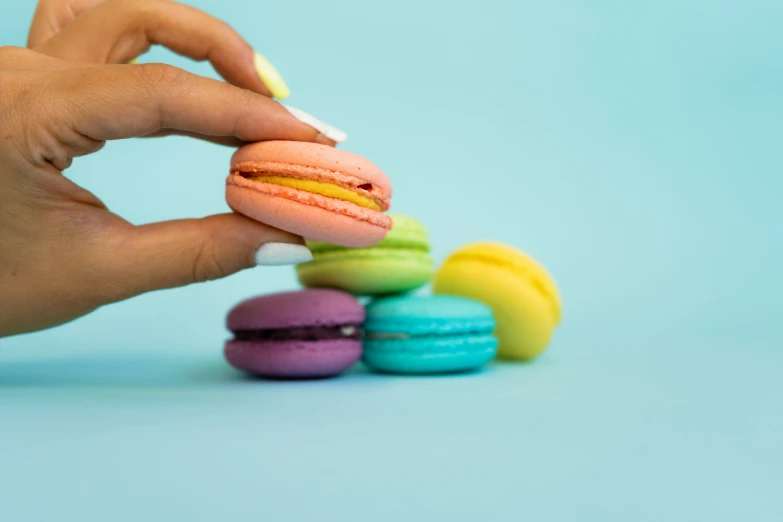 a hand holding a yellow and blue striped doughnut in front of a stack of rainbow colored macarons