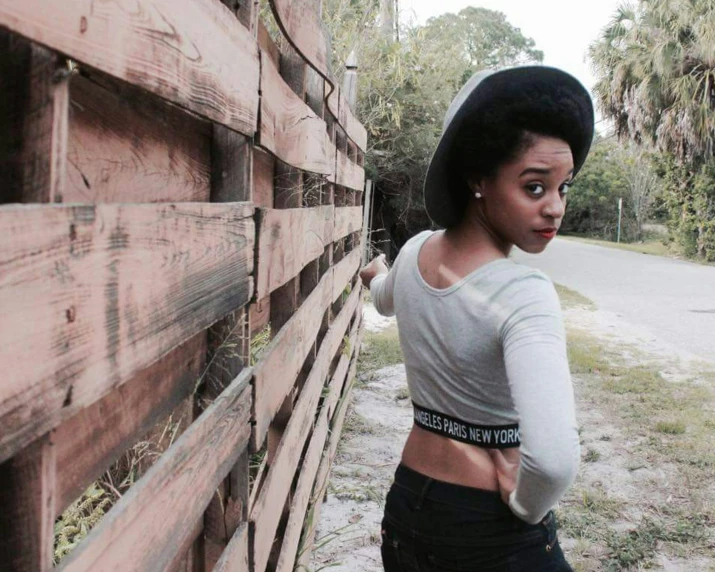a young black woman in high rise jeans and a hat leans against a wooden fence