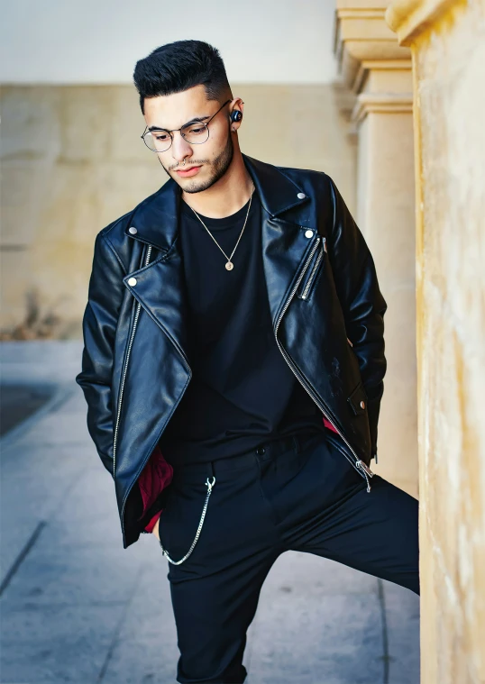 man with black hair in a leather jacket leaning against a column