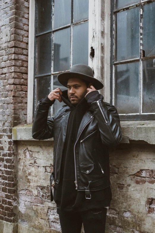 a man in a black hat and jacket leaning against a building