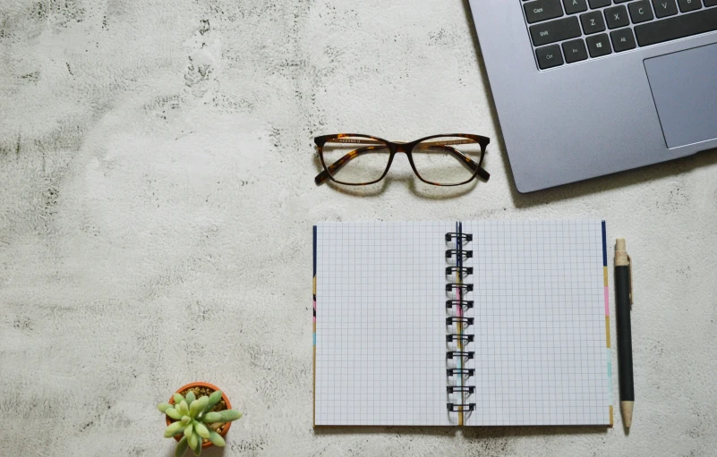 a notepad and glasses next to a laptop and a plant