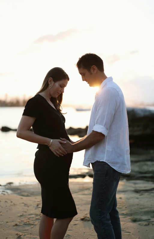 a pregnant couple standing on a beach with the sunset in the background