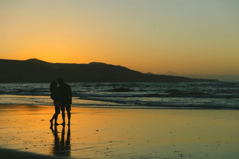 two people walk on the sand of an empty beach at sunset