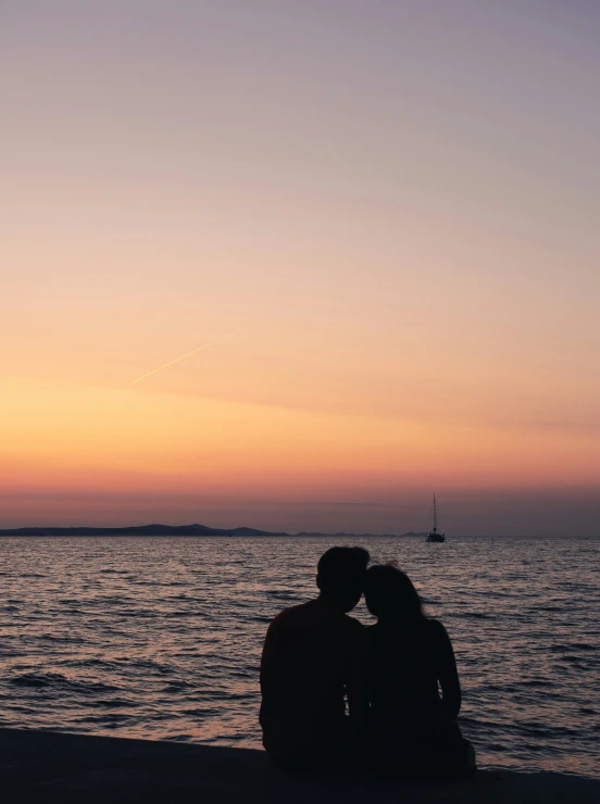 two people sitting at the waters edge in front of a boat