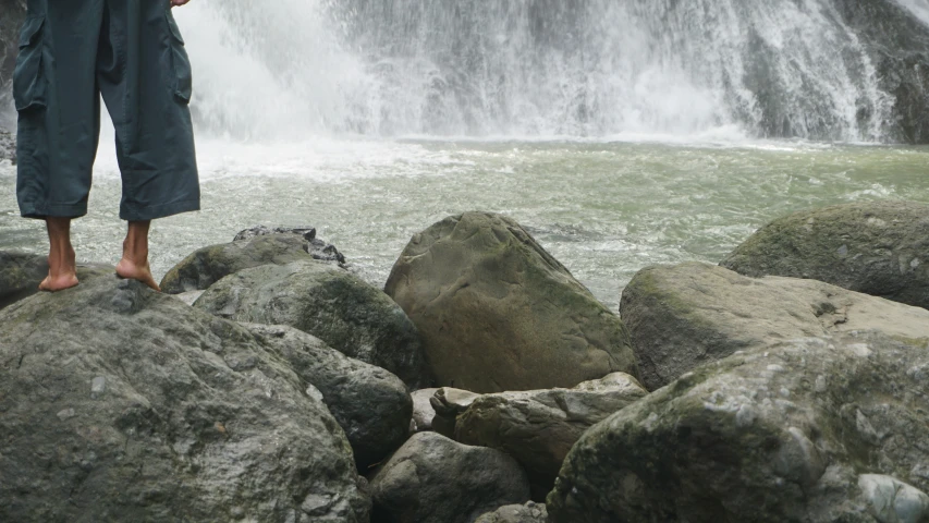 the legs of a person standing on large rocks near a waterfall