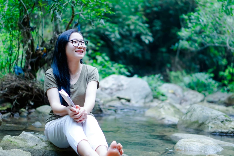 a woman with long black hair sitting on some rocks near a river