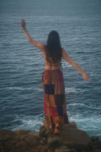 woman wearing a multi - color dress is standing on the rocks near water