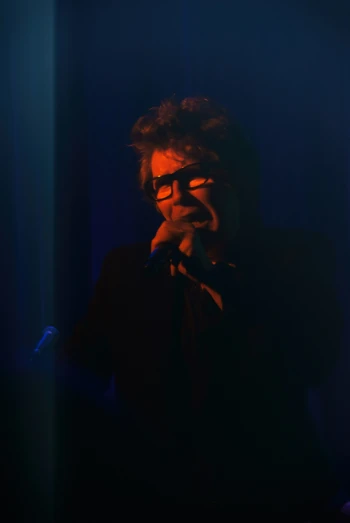 a man with glasses sings into microphone in dark area