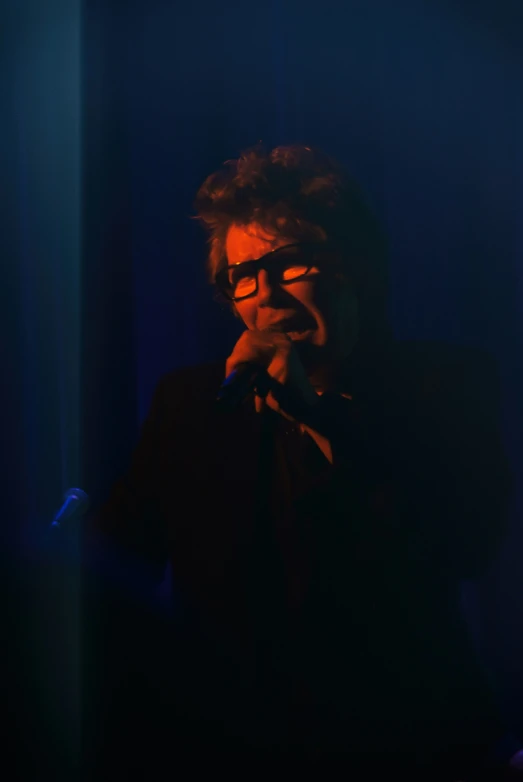 a man with glasses sings into microphone in dark area