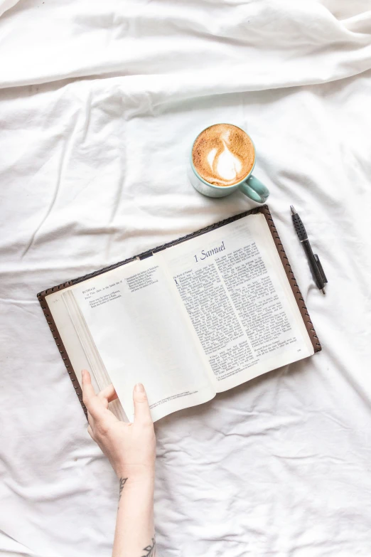 a person holding an open book with a cup of coffee and pen on top