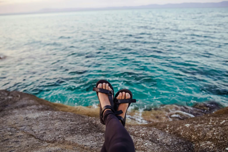 someones feet with shoes that have sandals on near the water