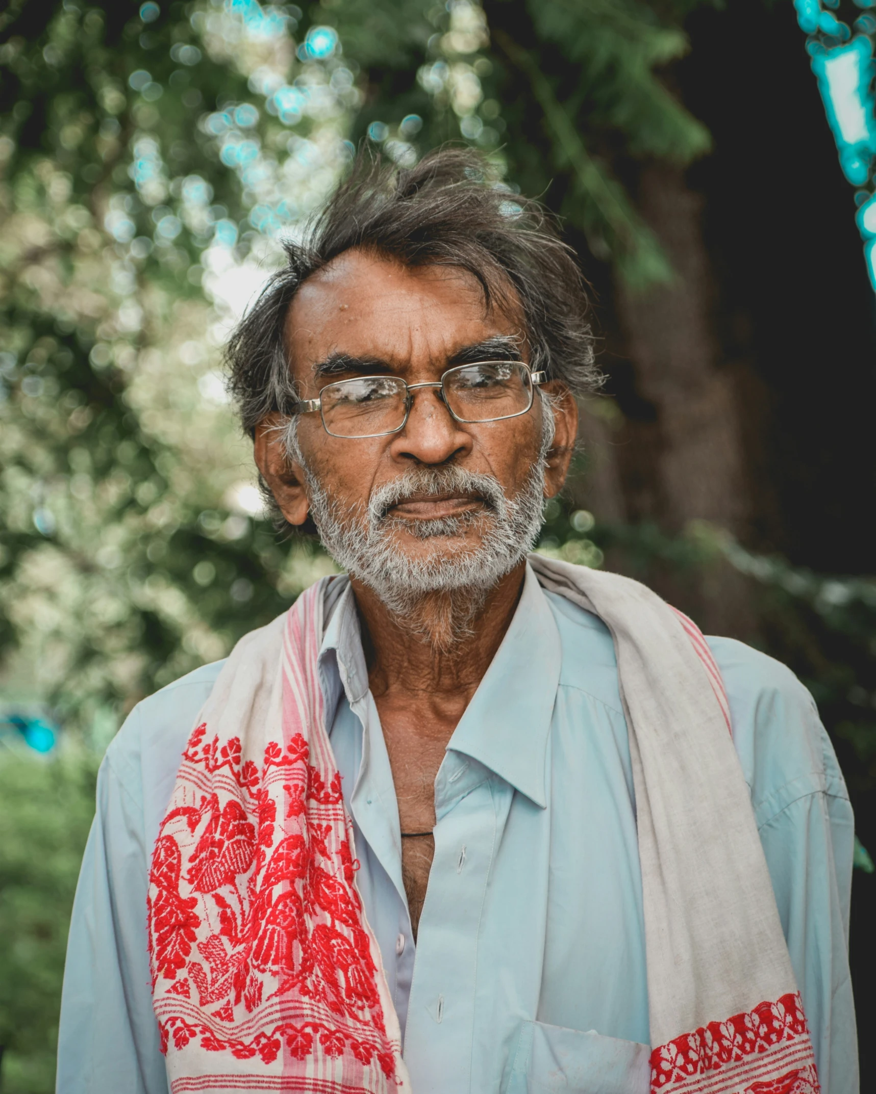 a man with glasses stands near trees, with a scarf on his shoulder