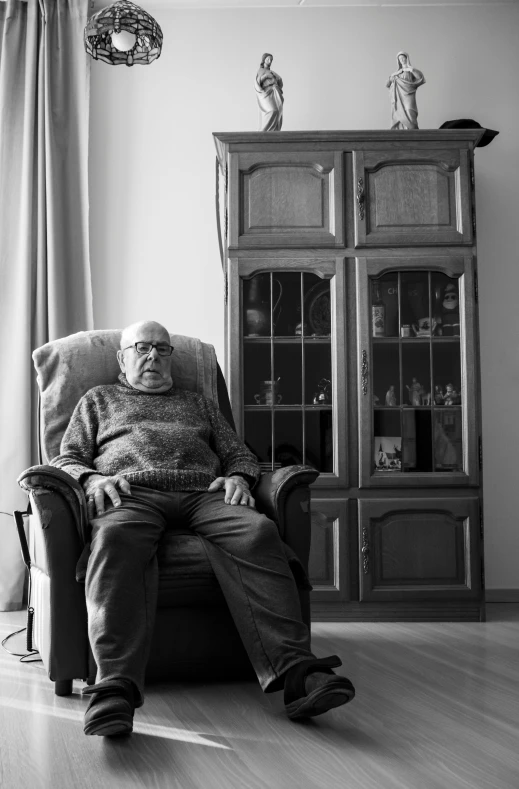 an old man sitting in a chair by a china cabinet