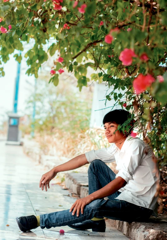 a boy in jeans sitting on the concrete next to a bush with flowers