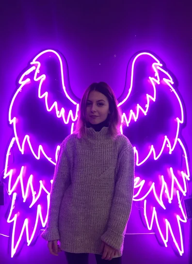 a woman is standing in front of a purple neon sign with wings