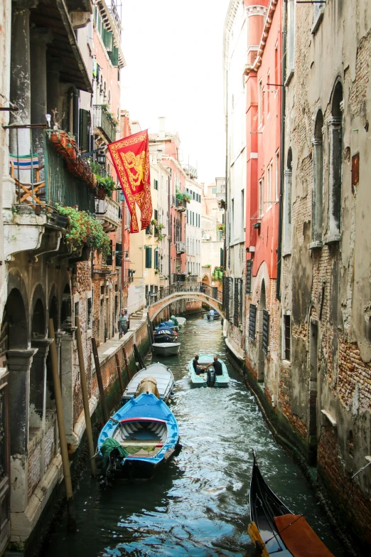 a narrow waterway with boats moored on both sides and red banners floating above the buildings