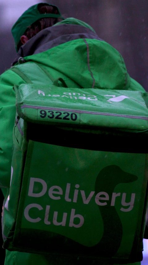 a man standing outside with his back to the camera and wearing a jacket that reads delivery club