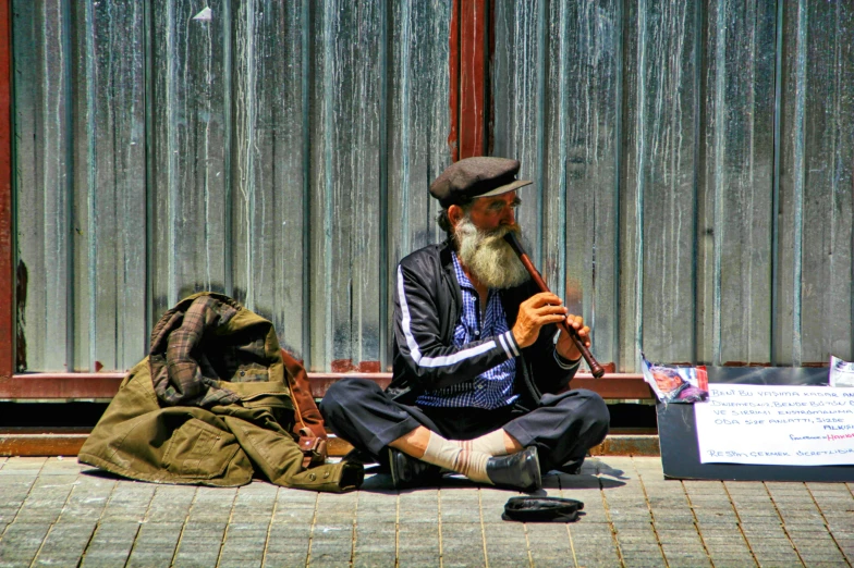 man with a long beard sits near a bag on the sidewalk