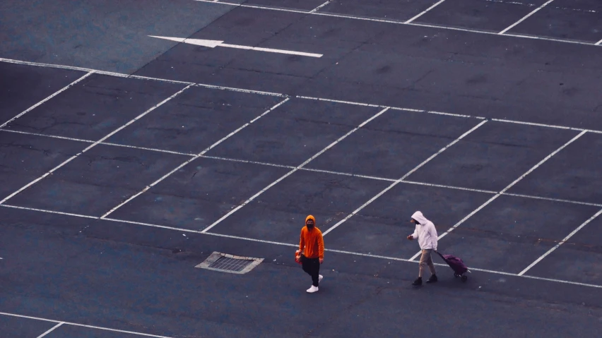 an aerial view of two people with luggage and one with an umbrella walking in the middle of a parking lot