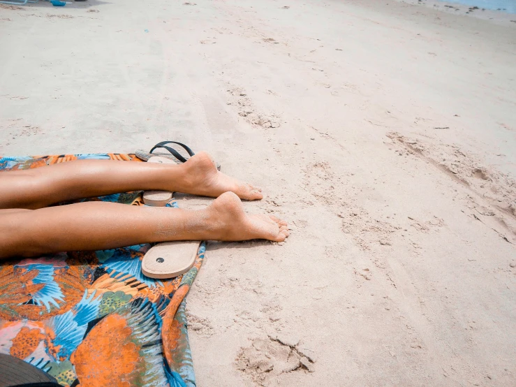 a woman's legs and toes on the sand near an orange flower blanket