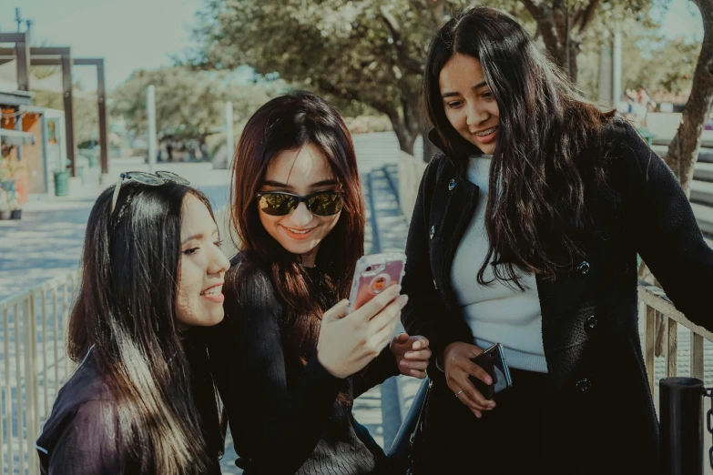 three young women stand in front of a tree and look at a cell phone