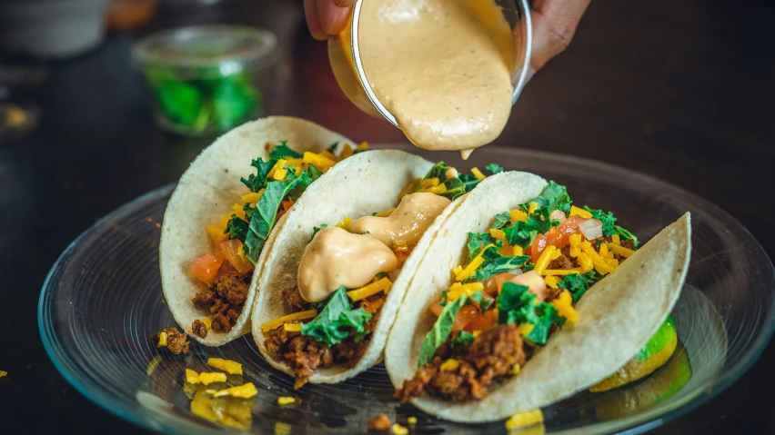 three tacos sit on a glass plate with sauce being poured over them