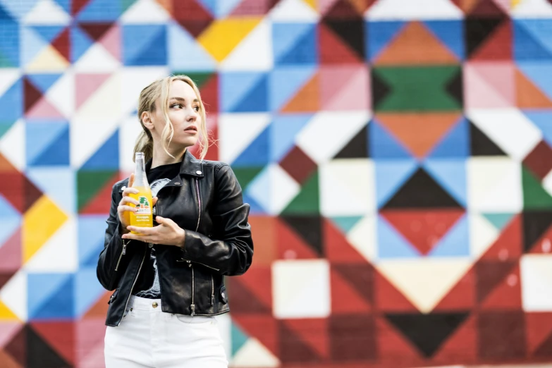 a woman in black jacket holding beverage next to a colorful wall