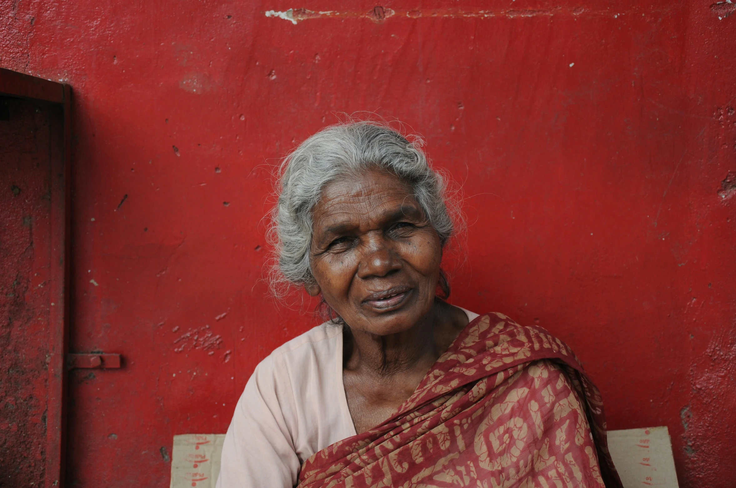 an elderly woman wearing a brown and red sari sitting outside a red wall