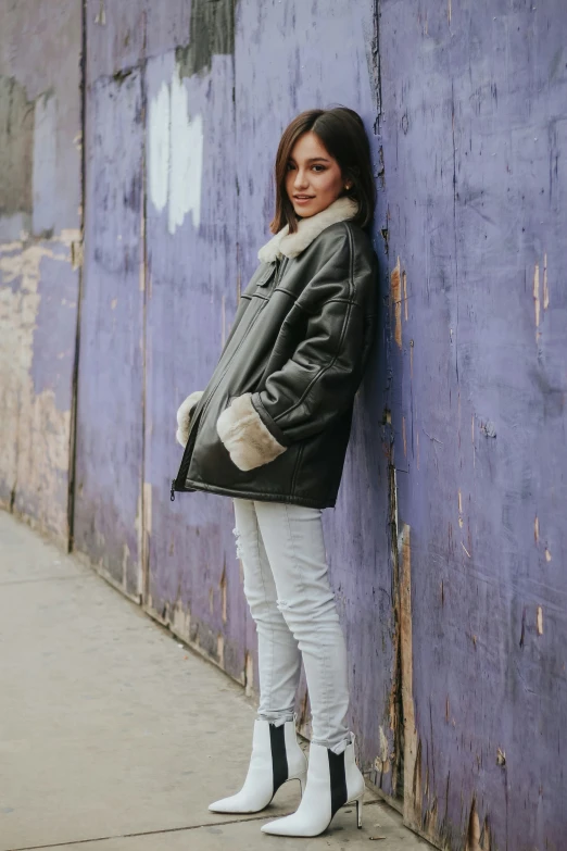 a young woman is standing in front of a wall wearing white pants and a leather jacket