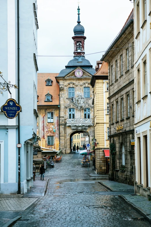 a cobblestone road has an archway on one side and a clock tower at the other