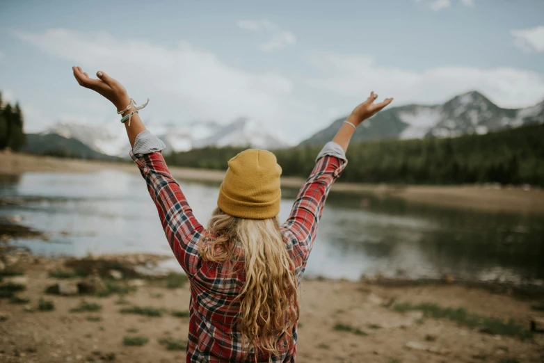 a girl in a plaid jacket and hat stands next to the water