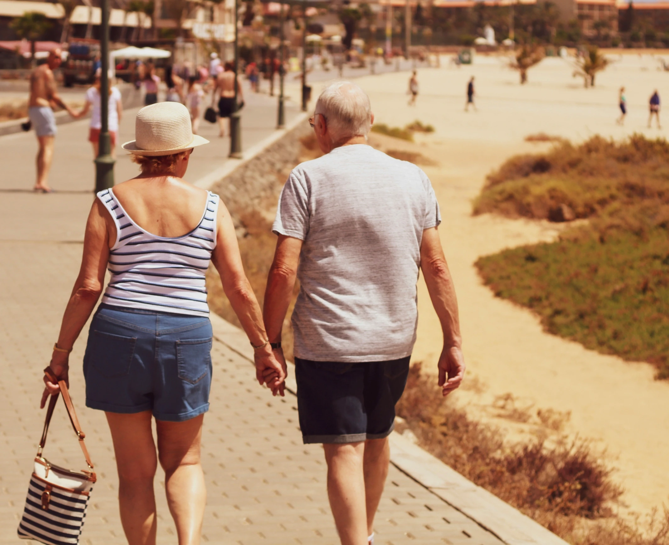 an older couple holds hands walking on the boardwalk at a beach