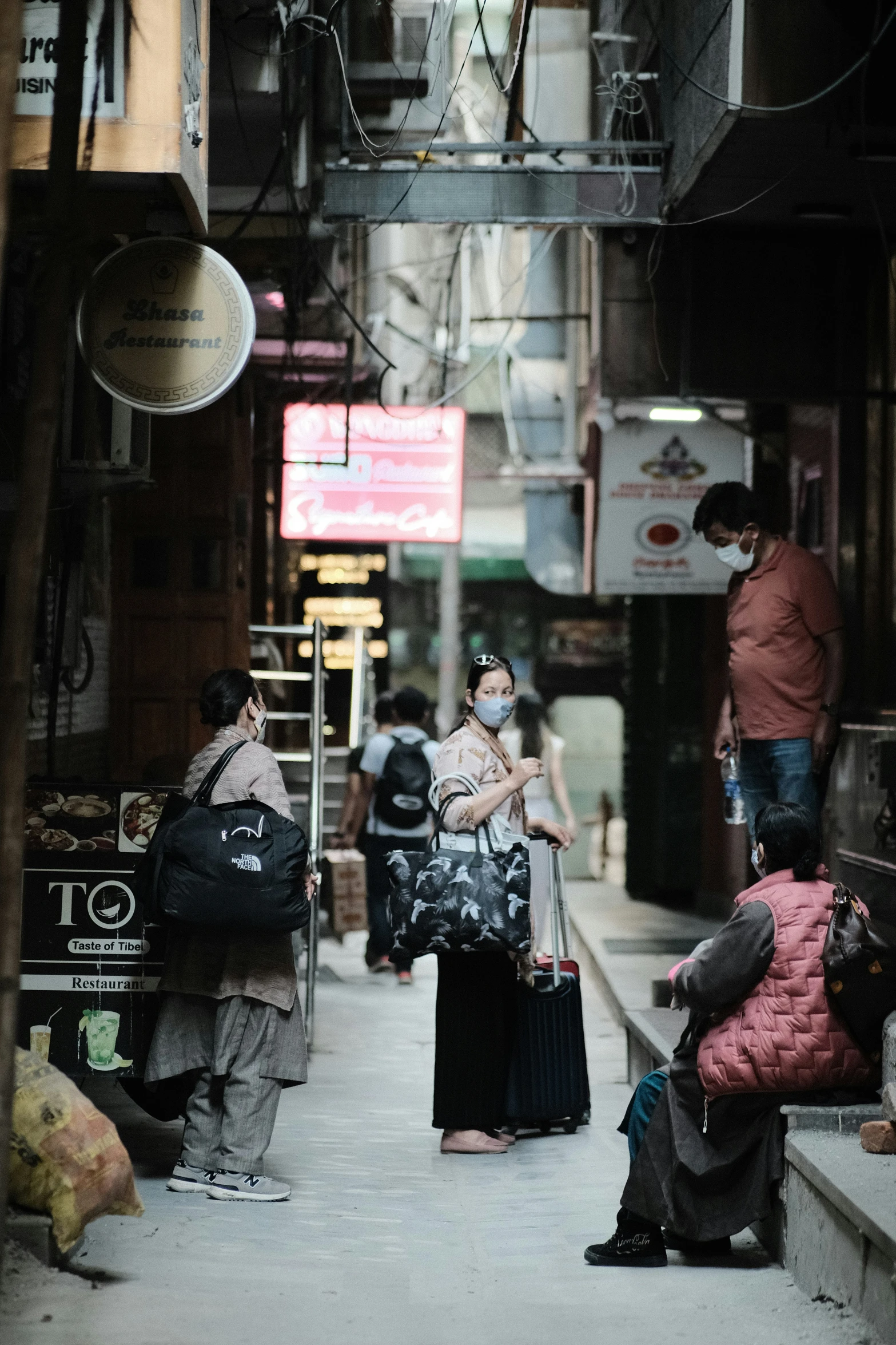 people with luggage walk down an alleyway while wearing masks