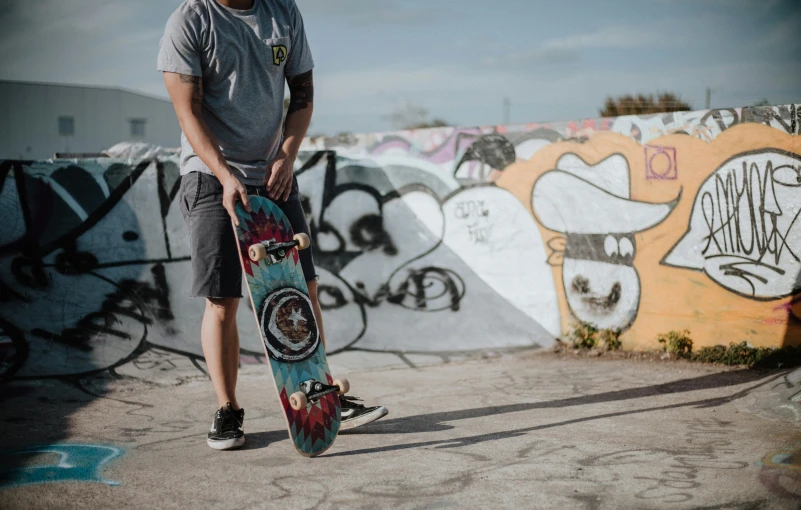 a guy holds his skateboard in front of a large wall covered with graffiti