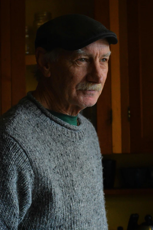 an older man standing by a kitchen stove with his hat turned to the side
