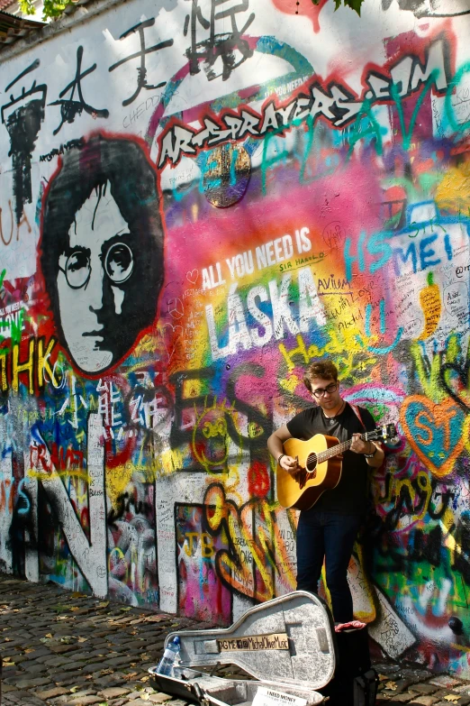 a man with a guitar poses near a wall covered in grafitti