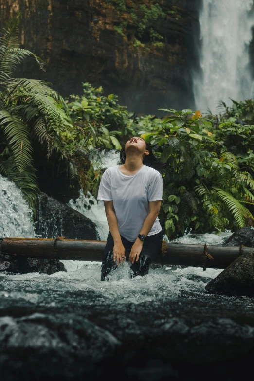 a man in a white shirt sitting on a log and watching a waterfall