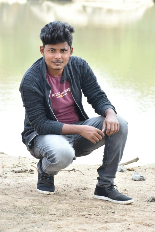 man with black hair sitting on a dirt next to water