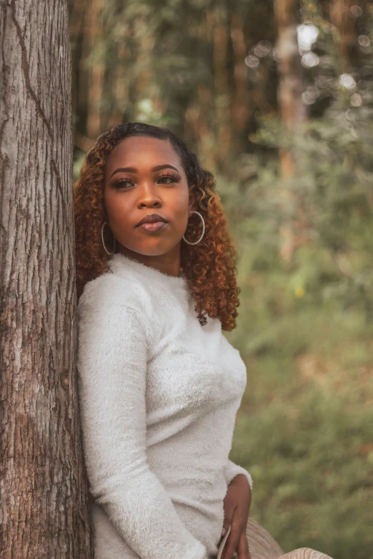 a young black woman stands by a tree, in a white sweater and beige skirt