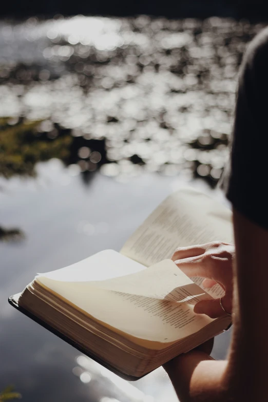 someone holding a book while sitting in front of a river