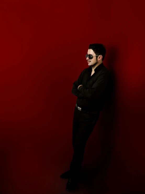 man in black suit leaning on red wall wearing sunglasses