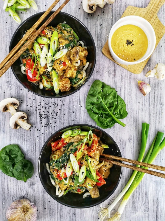 stir fryed vegetables in two black plates next to bowls with chopsticks and a mug of mustard