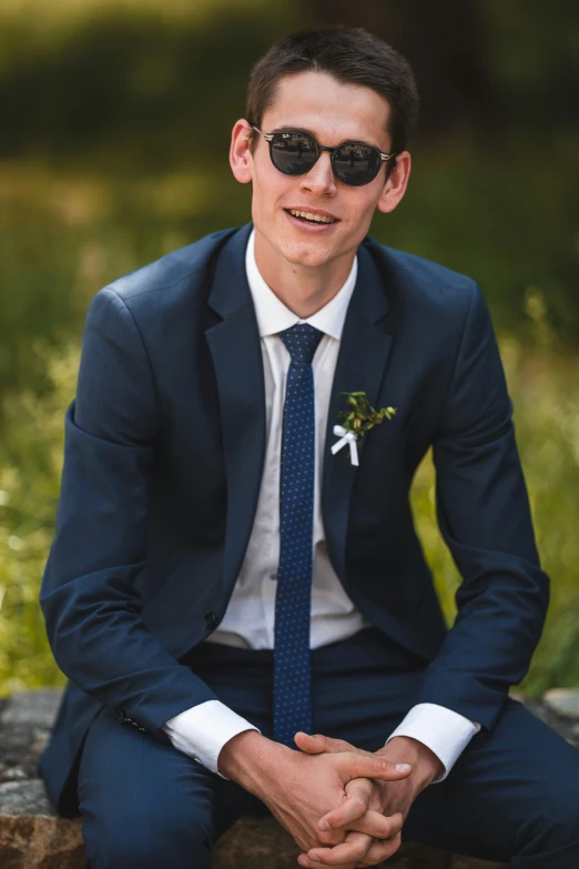 a man in glasses and suit with white flower sitting on a rock