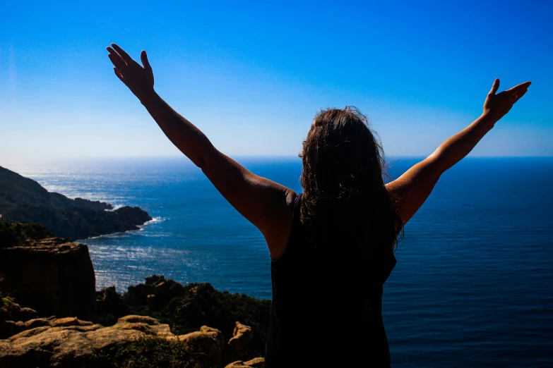 a woman with hands spread wide, arms up in front of her overlooking the ocean