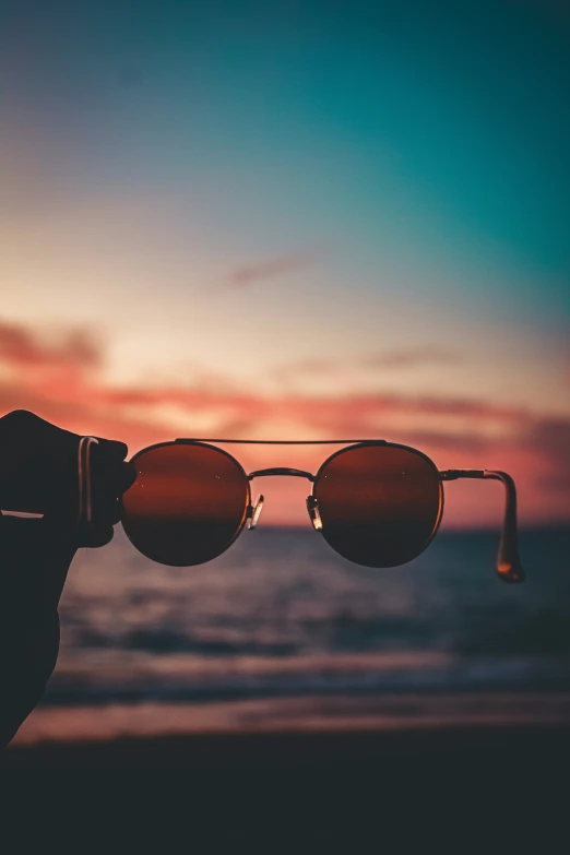 someone holding onto a pair of sunglasses with a sunset in the background