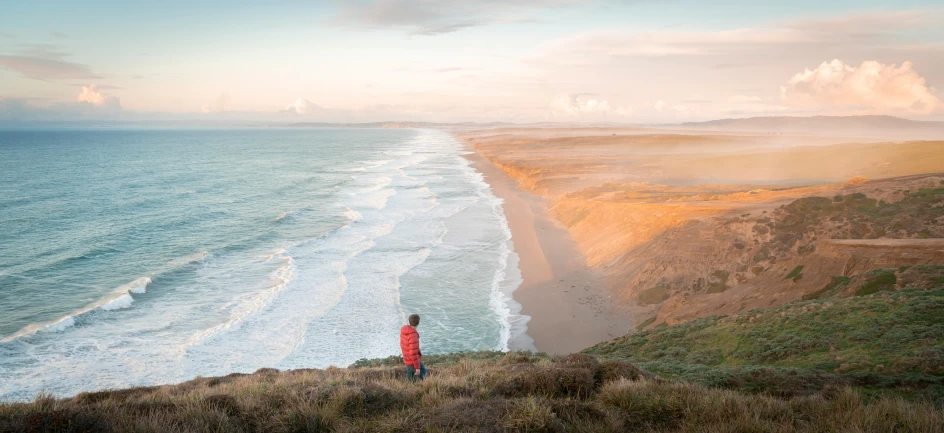 a lone person walks along the edge of a cliff overlooking the beach