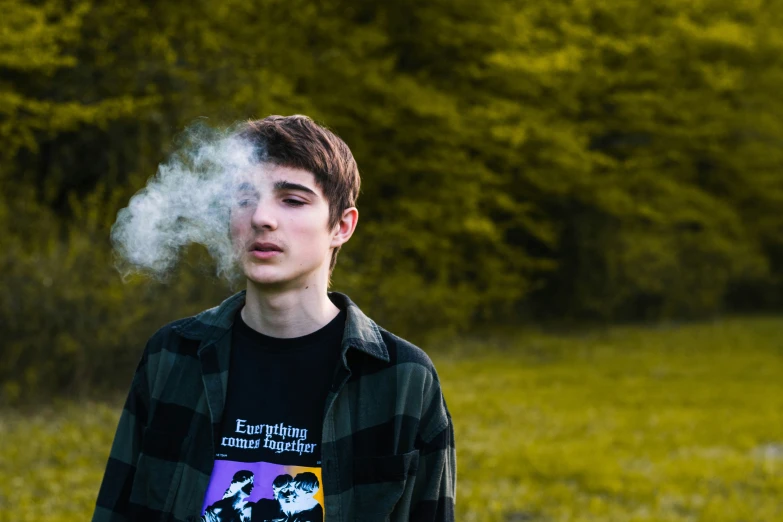 a young man is smoke by himself in front of some trees