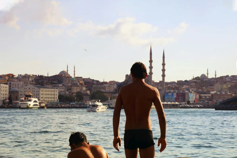 two young men standing near the water with a city in the background