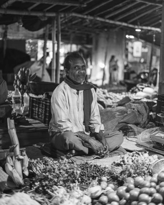 a black and white pograph of a man sitting on an outdoor produce stand